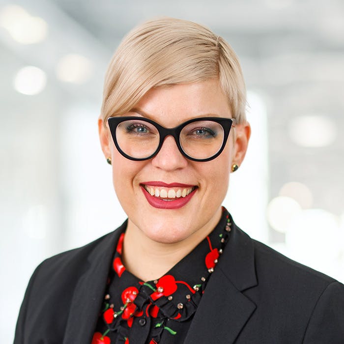 Lächelnde Frau mit blonden Haaren, schwarzer Brille und schwarzem Blazer.